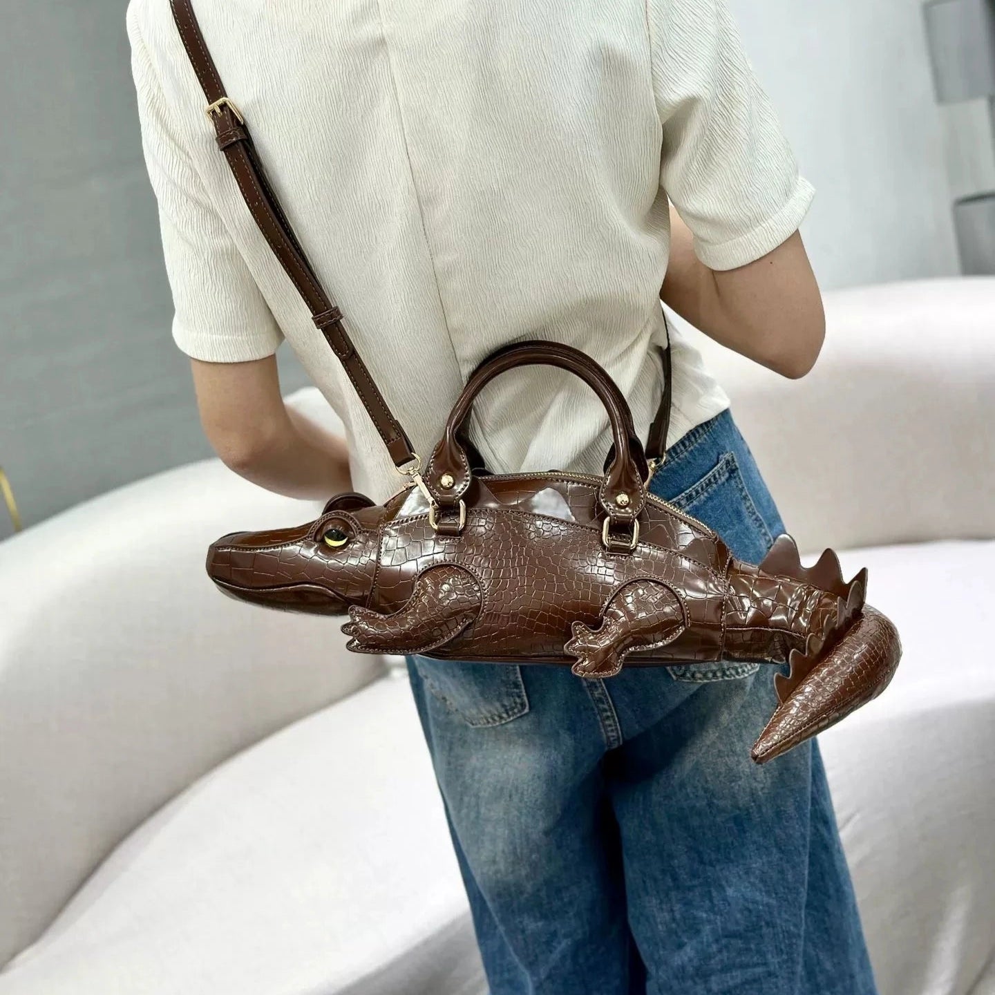 Person wearing a white shirt and blue jeans with a brown alligator-shaped handbag.